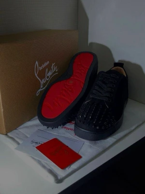 Christian Louboutin svarta sneakers med nitar - Svarta sneakers från Christian Louboutin med ikonisk röd sula och svarta nitar på tån. Skorna har snörning och rund tå, tillverkade i skinn med lyxig känsla. Kommer med originalkartong, dustbag och äkthetskort. Perfekt för dig som gillar edgy designerstil. Kvitto 🧾finns 