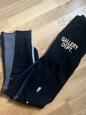Svarta mjukisbyxor Gallery Dept. - Säljer ett par svarta mjukisbyxor från Gallery Dept. med vit logotyp på benet. Byxorna har elastisk midja och färgstänk som detalj. Ena benet har grå och svarta paneler för en cool kontrast. Perfekt för chill eller streetwear.
