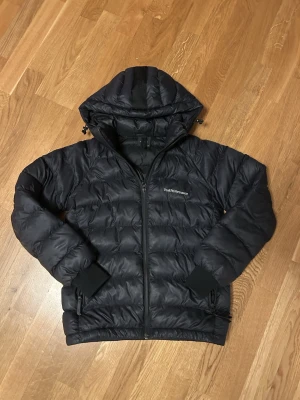 Svart dunjacka Peak Performance - Svart dunjacka från Peak Performance med huva och dragkedja framtill. Jackan har quiltad design, ribbade muddar vid ärmslut och två sidofickor med dragkedja. Perfekt för kalla dagar och har en stilren look med broderad logga på bröstet. Hör av er vid frågor! Passar mellan 155-165 typ.