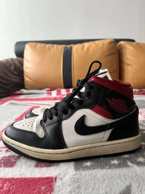 Nike Air Jordan 1 svart/vit/röd - Säljer ett par Nike Air Jordan 1 sneakers i klassisk svart, vit och röd färg. Skorna har högt skaft, snörning och rund tå. Materialet är skinn och de har en platt sula med tydligt Nike-mönster undertill. Perfekt för dig som gillar ikoniska sneakers med streetstyle-vibe.
