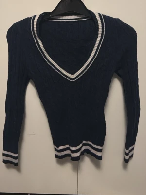 Mörkblå stickad v-ringad tröja - Snygg mörkblå stickad tröja från Crossbow med v-ringad hals och vita ränder vid halsringning, ärmslut och nederkant. Tröjan har långärmade och ribbade detaljer som ger en sportig vibe. Perfekt för dig som gillar klassisk stil med en twist.