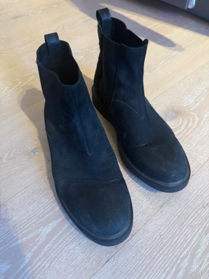 Svarta chelsea boots från Vagabond - Säljer ett par svarta chelsea boots från Vagabond i mocka med platt sula och rund tå. Skorna har klassisk resår på sidorna och dragflik bak för enkel påtagning. Perfekta för dig som gillar stilrena och tidlösa boots.