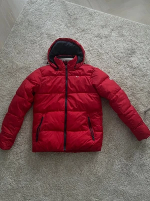 Röd pufferjacka med huva - Riktigt snygg röd pufferjacka från Tommy Hilfiger med svart dragkedja och två fickor med dragkedja. Jackan har en stor huva och quiltad design som ger extra volym och värme. Perfekt för kalla dagar och har en modern look som sticker ut. Säljer så billigt för behöver pengar