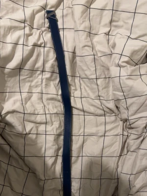 Blå och svart bälte med metallspänne - Säljer ett stilrent bälte i blått och svart med ett snyggt metallspänne. Bältet har vit kontrastsöm längs kanterna och är gjort i ett slätt material som ger en clean look. Perfekt accessoar för att lyfta din outfit.