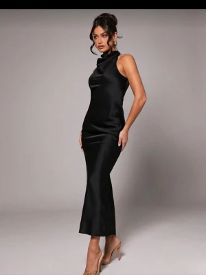 Svart långklänning med spetsdetalj - Säljer en elegant svart långklänning i satinliknande material med hög hals och ärmlös design. Baksidan har en snygg spetsdetalj och öppen rygg som ger en extra touch. Perfekt för dig som vill sticka ut på festen! Passformen är slim och klänningen har en stilren siluett.