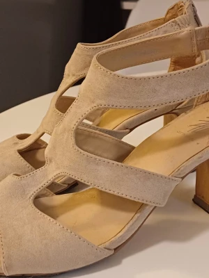 Beige mockasandaler med klack, Din Sko - Säljer ett par snygga sandaler från Din Sko i beige mocka med bred klack och öppna partier. Skorna har rund tå och dragkedja bak för enkel påtagning. Perfekt för dig som gillar stilrena och bekväma klackar.