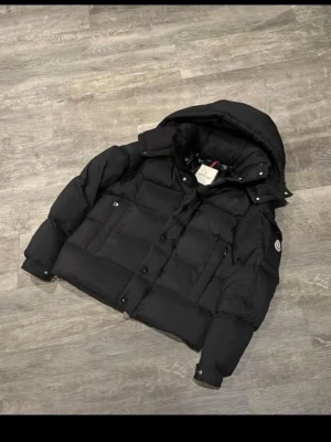 Svart pufferjacka från Moncler - Säljer en svart pufferjacka från Moncler med huva och logotyp på ärmen. Jackan har en quiltad design, dragkedja och knappar framtill samt praktiska fickor. Perfekt för kalla dagar och har en riktigt schysst streetstil.