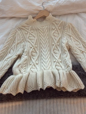 Krämvit stickad tröja från Zara - Superfin krämvit stickad tröja från Zara med snygga flätmönster och små bollar i strukturen. Tröjan har långärmade ärmar, hög hals med knappstängning och en volangkant nedtill som ger en extra touch. Perfekt för dig som gillar mysiga och trendiga plagg.