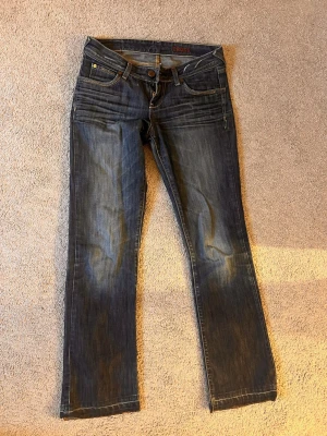 Mörkblå jeans från Cross - Säljer ett par jättesnygga mörkblå jeans från Cross med klassisk femficksdesign och snygga detaljer på bakfickorna. Jeansen har raka ben och låg midja, med slitningar och kontrastsömmar. Knappen man stänger byxorna med är lös men lossnar inte när byxorna sitter på. Har även en reva i ena byxbenet som visas.  