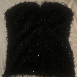 Svart paljettkorsett från Zara - Säljer en svart top från Zara med glittriga paljetter och fluffig textur. Toppen har knappar framtill och en v-ringning i fram.