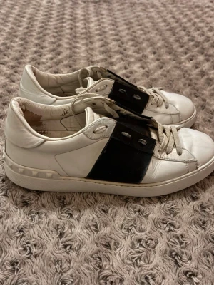 Valentino Open - Snygga vita och svarta sneakers från Valentino Garavani med bred svart läderdetalj över snörningen och klassiska nitar på hälen. Skorna har rund tå, platt sula och är tillverkade i skinn. Perfekta för dig som gillar exklusiva och stilrena sneakers. De kommer utan dustbag och original boxen men de är äkta.