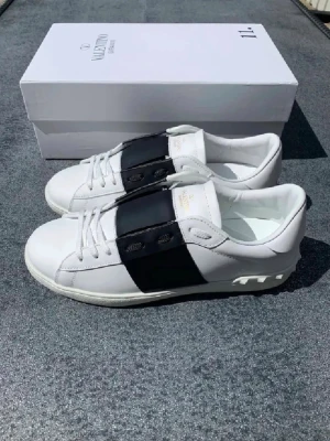 Valentino Garavani Open sneakers vit/svart - Snygga Valentino Garavani Open sneakers i vitt skinn med bred svart läderpanel över mitten. Klassisk rund tå, platt sula och vita snören. Märkeslogga i guld på plösen. Kommer med originalkartong, dustbag och tags. Perfekt för dig som gillar stilrena designer.  priset kan dikuteras 