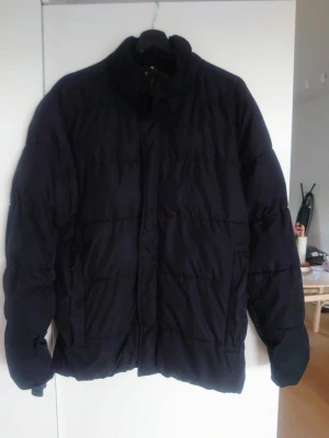 Svart pufferjacka från Dressmann - Svart pufferjacka från Dressmann i storlek M. Jackan har en hög krage, quiltad design och stängs med dragkedja. Perfekt för kalla dagar och har en clean look som funkar till många stilar. Materialet är polyester och jackan har långa ärmar.