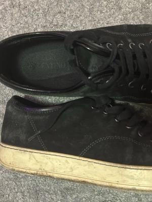 Svarta sneakers från Lanvin i mocka storlek 42 - Säljer ett par svarta sneakers från Lanvin med ovandel i mocka och detaljer i blankt skinn. Skorna har vita, slitna gummisulor och svarta skosnören. Klassisk låg modell med rund tå och kontrastsömmar. Perfekta för dig som gillar stilrena och exklusiva sneakers.