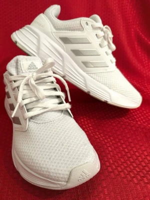 Vita Adidas Cloudfoam sneakers 39 - Säljer ett par fräscha vita Adidas Cloudfoam sneakers med silvriga detaljer och klassiska tre ränder på sidan. Skorna har snörning, rund tå och platt sula. Ovandelen är i mesh och syntet för extra komfort. Perfekta för dig som gillar stilrena och sportiga sneakers. Textil 