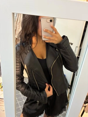 Svart skinnjacka med dragkedjor - Säljer en svart skinnjacka med coola detaljer som flera silvriga dragkedjor och quiltade partier på axlar och ärmar. Jackan har en figurnära passform och snygga sömmar som ger en edgy look. Lite skrapor som knappt syns annars inga defekter. 