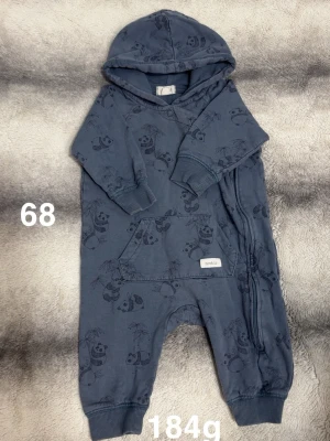 Blå overall med panda-mönster från Newbie - Mysig blå overall från Newbie med huva och sött pandamönster. Overallen har en stor ficka framtill, muddar vid ärm- och benslut samt dragkedja längs sidan för enkel påklädning. Perfekt för små äventyr och lek.🌸 Fint använt skick. Storlek 68