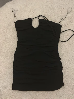 Svart kort klänning från Zara - Säljer en svart, figurnära kort klänning från Zara i storlek XS. Klänningen har tunna axelband, snygg rynkad design och en liten v-ringning framtill. Perfekt för dig som gillar stilrena och trendiga plagg med enkel men cool look.