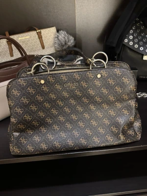 Svart handväska från Guess - Säljer en snygg svart handväska från Guess med brunt monogrammönster och silvriga metalldetaljer. Väskan har flera fack med dragkedja och praktiska handtag i samma mönstrade material. Perfekt för dig som gillar stilrena accessoarer med lyxig känsla.