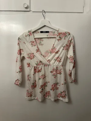 Blommig omlottopp från Bik Bok - Supersöt omlottopp från Bik Bok i vitt med rosa blommigt mönster. Toppen har v-ringning, trekvartsärm och är gjord i mjuk bomull. Perfekt för dig som gillar romantisk och feminin stil.