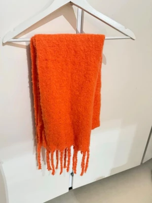 Orange stickad halsduk med fransar - En riktigt snygg orange stickad halsduk med långa fransar i ändarna. Halsduken är mjuk och fluffig, perfekt för att ge lite extra färg till din outfit. Enkel design utan mönster och passar till alla tillfällen när du vill hålla dig varm.
