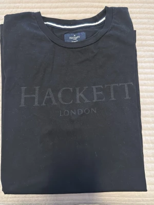Svart t-shirt från Hackett London - Snygg svart t-shirt från Hackett London med diskret logotyp tryckt på bröstet. Klassisk rund hals och korta ärmar, perfekt för en clean och stilren look. Tillverkad i mjuk bomull som känns skön mot huden.