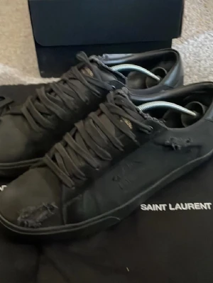 Svarta sneakers från Saint Laurent - Säljer ett par svarta Saint Laurent sneakers i skinn med slitna detaljer och snörning. Klassisk låg modell med rund tå och diskret logga på plösen. Snygg minimalistisk look med råa kanter och ton-i-ton sula. Perfekta för dig som gillar streetstyle.