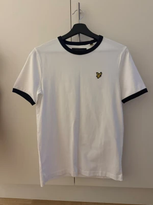 Vit t-shirt från Lyle & Scott - Vit t-shirt från Lyle & Scott med svart ribbad krage och svarta muddar vid ärmslut. Klassisk rund hals och korta ärmar. På bröstet finns den ikoniska gula fågeln broderad. Tillverkad i mjuk bomull som känns skön mot huden.