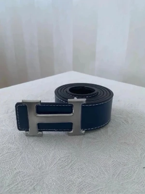 Blå skinnväst med silverdetaljer - Snygg blå väst med markerade vita sömmar och en stor silvrig H-detalj framtill. Västen har en rak och klassisk form, tillverkad i skinn med en stilren look. Perfekt för dig som vill sticka ut med en cool accessoar.