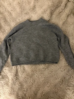 Grå stickad tröja med rund hals - Mysig grå stickad tröja med rund hals och långa ärmar. Tröjan har en enkel design med ribbade muddar vid ärmslut och nederkant. Perfekt för att hålla sig varm under kyliga dagar och passar till jeans eller kjol.