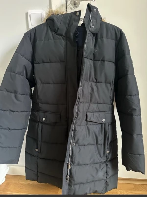 Svart pufferjacka från Five Seasons - Svart pufferjacka från Five Seasons med huva och fuskpälsdetalj. Jackan har två stora fickor med lock och knappar, samt dragkedja framtill. Tillverkad i 100% polyester, perfekt för kalla dagar. Designad i Sverige, stilren och praktisk. Endast använd en gång :) 