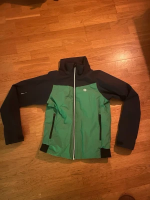 Grön och mörkblå vindjacka 8848 Altitude - Snygg vindjacka från 8848 Altitude i grön och mörkblå färg med vit dragkedja. Jackan har hög krage, två sidofickor med svarta dragkedjor och sportig design. Perfekt för dig som gillar att vara aktiv utomhus och vill ha en cool look.