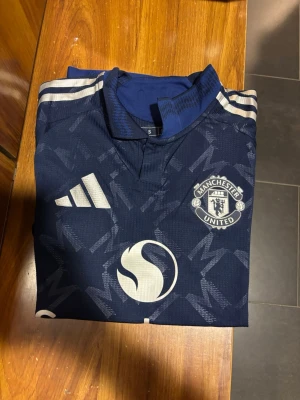 Manchester United fotbollströja - Snygg blå Manchester United tröja från adidas i slim fit. Materialet är lätt polyester och perfekt för fotboll eller träning. Officiell licensierad produkt med Snapdragon sponsor. Passar folk mellan 170-185. Slim fit! Om önskas så kan jag skicka bild på hur den ser ut på mig som är 184cm. Tveka inte att höra av er vid frågor eller funderingar 🤗