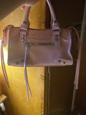 Rosa handväska med dragkedja - Säljer en snygg rosa handväska i skinnimitation med coola metalldetaljer och dragkedja framtill. Väskan har två handtag och dekorativa remmar på sidorna. Perfekt storlek för det viktigaste och ger en trendig touch till din outfit.