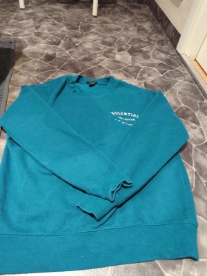 grön sweatshirt från Amisu - grön sweatshirt från Amisu med tryck 'Essential Collection' på bröstet. Tröjan har rund hals, långa ärmar och ribbade muddar vid ärmslut och nederkant. 