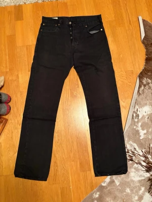 Svarta Levi's 501 jeans W33 L34 - Svarta Levi's 501 jeans med klassisk rak passform och knappgylf. Jeansen har fem fickor, silverfärgade knappar och detaljer i mässing. Materialet är slitstark denim i bomull. Perfekt för dig som gillar tidlös stil och vill ha ett par jeans som funkar till allt.