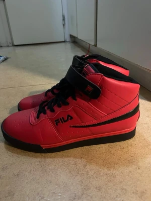 Röda high top sneakers från FILA - Säljer ett par röda high top sneakers från FILA med svarta detaljer och snörning. Skorna har kardborreband upptill, svart sula och är tillverkade i syntetmaterial. Perfekta för dig som vill sticka ut med en sportig och retro vibe.