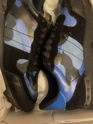 Valentino Rockrunner blå sneakers - Säljer ett par Valentino Rockrunner sneakers med blått och svart camouflage-mönster. Skorna har svarta snören, platt sula och detaljer i både mocka och skinn. Metallplatta med Valentino Garavani-logga på plösen. Riktigt snygga och unika sneakers för dig som gillar statement pieces.
