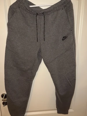 Grå mjukisbyxor Nike tech, storlek M - Säljer ett par grå mjukisbyxor från Nike i storlek M. Byxorna har snörning i midjan, ribbade muddar vid bensluten och praktiska fickor. Materialet är mjukt och bekvämt, perfekt för chill eller träning. Nike-logga på vänster ben och snygga detaljer vid fickorna.