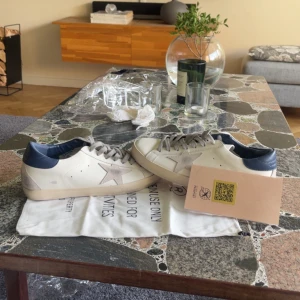 Golden Goose sneakers med blå detalj 42 - Snygga sneakers från Golden Goose i vitt skinn med grå mockadetaljer och stjärna på sidan. Baktill finns en mörkblå hälkappa i skinn och grå snörning. Klassisk platt sula och rund tå. Perfekta för dig som gillar stilrena men ändå unika skor.