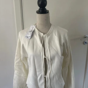 Vit stickad kofta  med snörning - Supersnygg vit stickad kofta från Gina i storlek S. Koftan har långa ärmar och cool snörning framtill som ger en unik look. Mjuk och skön kvalitet, perfekt för dig som gillar stilrena och trendiga plagg. Helt ny med prislapp 