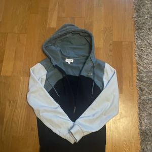 Lacoste hoodie i blått  - Snygg hoodie från Lacoste i färgblock med mörkblå kropp, ljusblå ärmar och gråblå axelparti. Klassisk krokodillogga på bröstet, huva med snörning och hel dragkedja framtill. Perfekt för chill dagar eller när du vill ha en sportig look. Köpt för 1600kr men säljer nu för 900kr