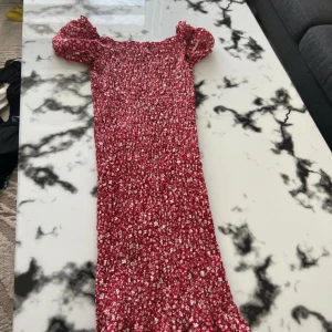 Röd blommig långklänning med puffärm - Säljer en röd långklänning med små vita blommor och puffärmar. Klänningen har en smockad, figurnära passform och offshoulder-design som ger en trendig vibe. Perfekt för dig som gillar romantiska detaljer och vill sticka ut med färg och mönster.