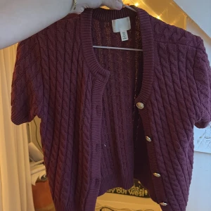 Kortärmad vinröd kofta med knappar - Säljer en vinröd kabelstickad kofta från H&M med korta ärmar och snygga guldfärgade knappar framtill. Koftan har ribbade kanter och ett klassiskt kabelmönster som ger en trendig look. Perfekt att slänga över en topp för extra stil.