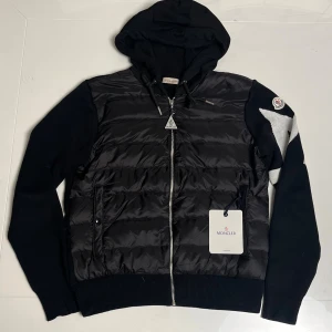 Svart pufferjacka med huva från Moncler / Cardigan  - Svart pufferjacka från Moncler med stickade ärmar och stor vit M-detalj på ena ärmen. Jackan har huva, dragkedja framtill och två fickor. Framsidan är quiltad och glansig medan baksidan och ärmarna är stickade. Ikonisk Moncler-logga på ärmen.