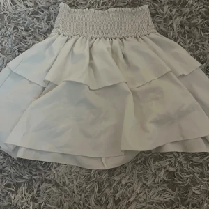 Vit volangkjol med smock - Supersöt vit kortkjol med bred smock i midjan och dubbla volanger. Kjolen har en luftig känsla och passar perfekt till varmare dagar. Den elastiska midjan gör den bekväm och enkel att styla med olika toppar.