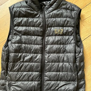 Svart dunväst från Emporio Armani EA7 - Svart quiltad dunväst från Emporio Armani EA7 med guldfärgad logga på bröstet. Västen har hög krage, dragkedja framtill och en stilren, sportig look. Perfekt för lager-på-lager och enkel att matcha med andra plagg.