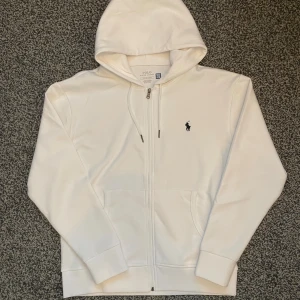 Vit zip hoodie från Polo Ralph Lauren - Säljer en vit zip hoodie från Polo Ralph Lauren med dragkedja och huva. Tröjan är i storlek S men sitter mer som M. Tröjan har det klassiska broderade logotypen på bröstet, ribbade muddar och fickor framtill. Perfekt för en clean och avslappnad look. Om du har några funderingar så är det bara att höra av.