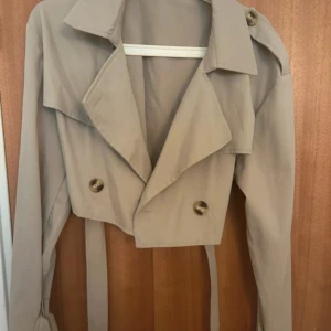 Beige croppad trenchcoat med bälte - Snygg croppad trenchcoat i beige med bred krage, knappar framtill och långa ärmar. Jackan har bälte som knyts i midjan och axelklaffar för extra stilpoäng. Perfekt för dig som gillar trendiga och unika jackor.