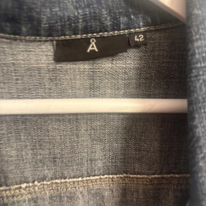 Klassisk blå jeansjacka från Å - Snygg blå jeansjacka från Åhléns i klassisk modell med silverfärgade knappar och två bröstfickor. Jackan har långa ärmar, krage och tydliga sömmar som ger en tidlös denimlook. Perfekt att slänga på sig över en hoodie eller t-shirt. Funkar bra som en större jacka till mini kläning. Pris går att diskuteras 